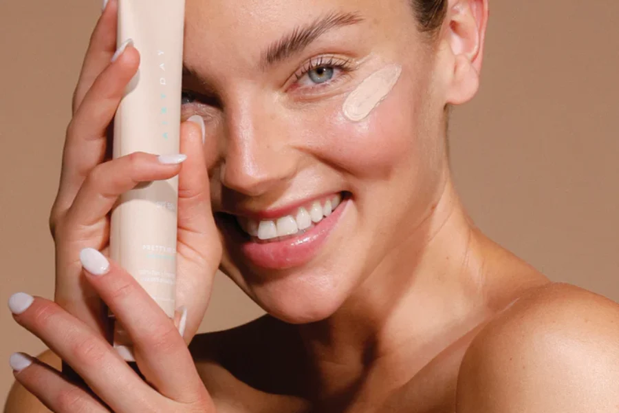 Smiling young woman holds tube of Airyday Sun Protection against her face. She has a sample of the sunscreen on her cheek.